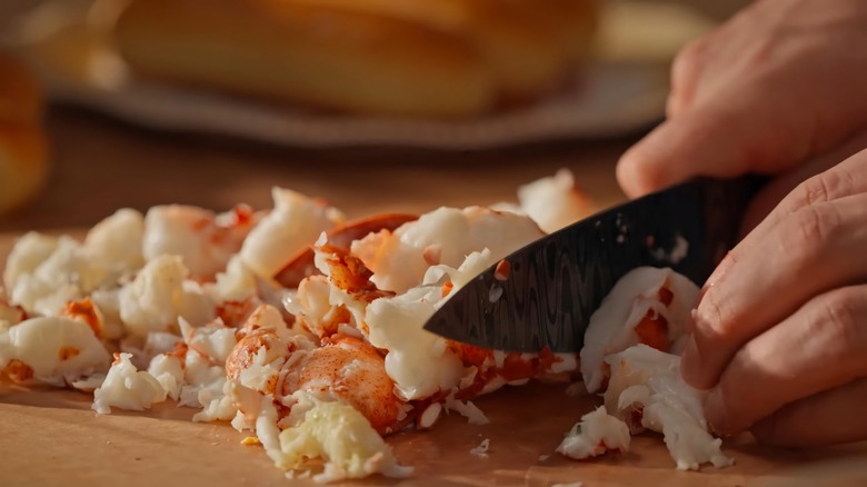 A close-up shot shows hands chopping cooked lobster meat into smaller pieces on a wooden cutting board with a black knife.