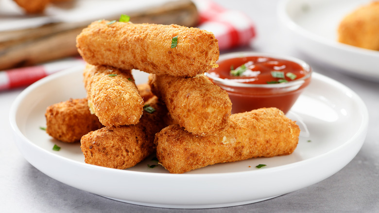 A stack of breaded and fried mozzarella sticks on a white plate with a red dipping sauce.