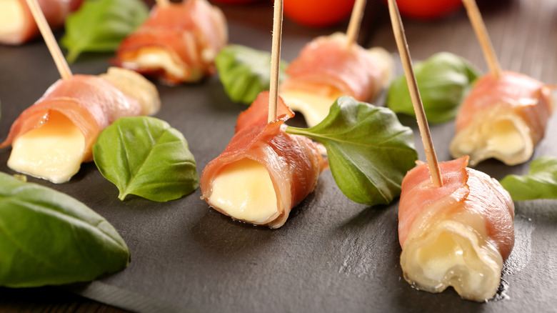 Prosciutto wrapped around a gooey white cheese and held together with a toothpick on a black platter with fresh basil leaves.