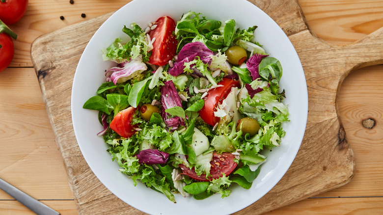 Salad with mixed greens, olives, and sliced tomato in a white ceramic bowl on a rustic wooden cutting board.