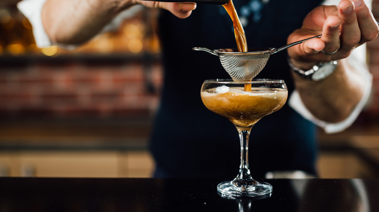 A close-up of a bartender preparing an espresso martini by pouring liquid through strainer into glass.