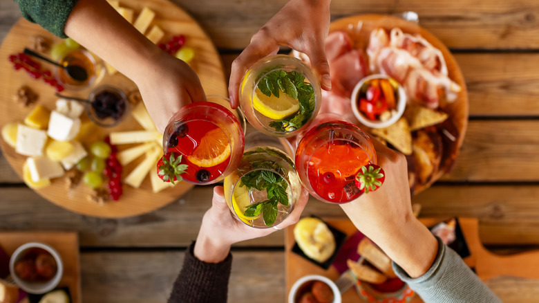 A birds eye view of four hands holding cocktails, clinking glasses. Below two charcuterie boards sit on a wooden table.