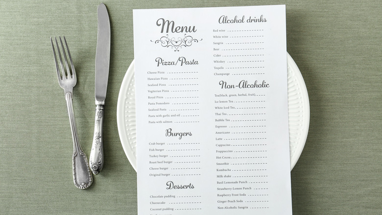 A white paper menu rests on a plate, a metal fork and knife to the left of it. The table cloth behind is light green.
