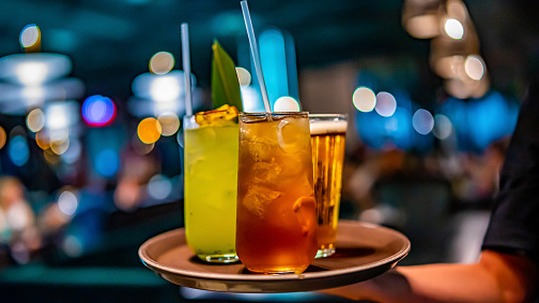 Three cocktails rest on a metal serving platter, a hand holding the bottom. The background is blurred.