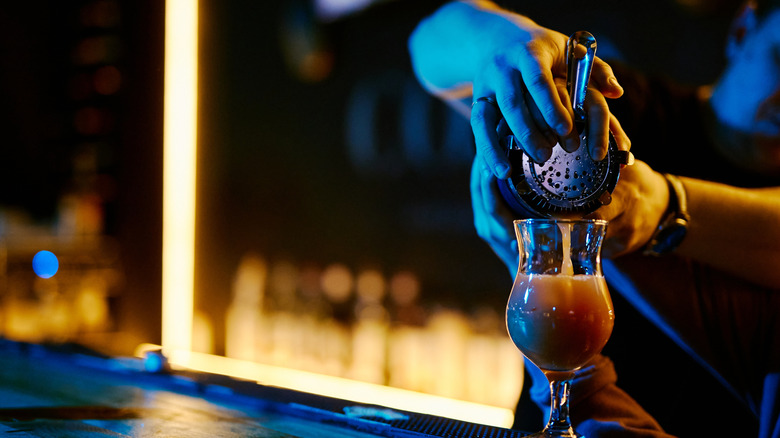 One bartender teaches another how to strain a drink into a glass in a darkened bar.
