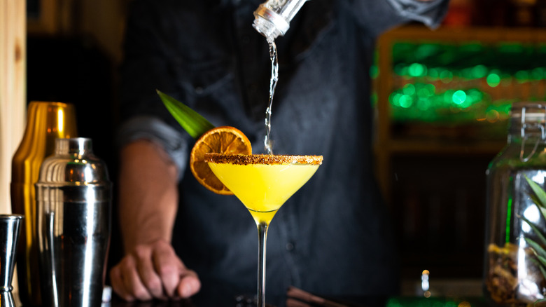 A bartender in the background pours alcohol into a yellow drink on a bar.