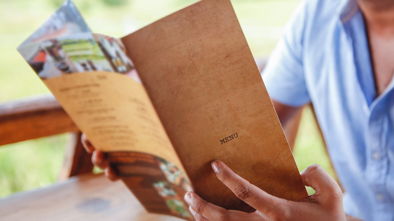 A man holds an unfolded paper menu, a wooden table and green grass in the background. He is wearing a blue shirt.