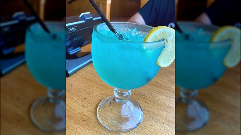 A bright blue liquid in a goblet style glass garnished with a wheel of lemon