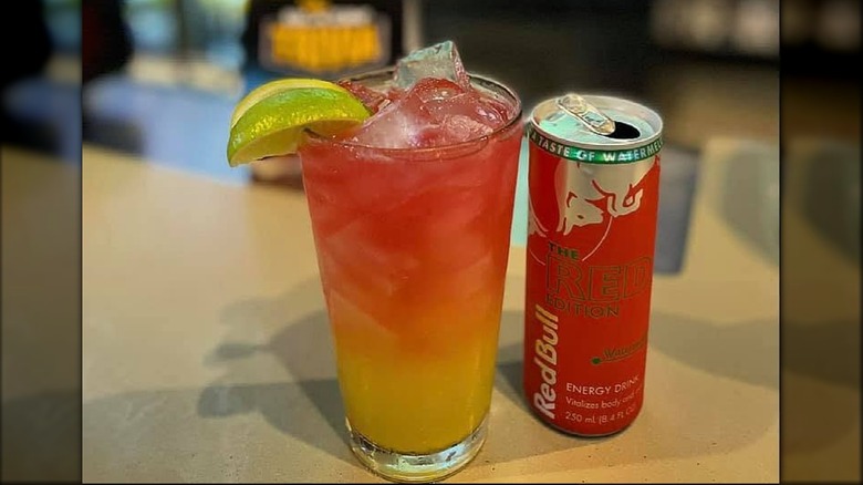 A pint glass with a layered orange and red liquid and a lime wedge next to a can of Red Bull