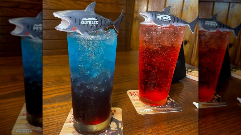 Two tall glasses on a wooden table filled with colorful liquids and topped with a plastic shark
