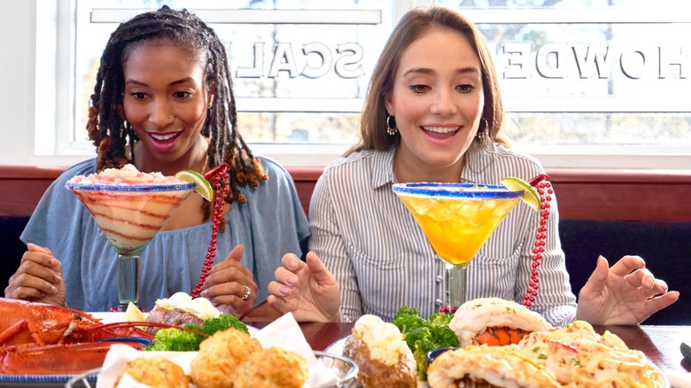 Two women smile and stare at oversized margaritas on the table in front of them