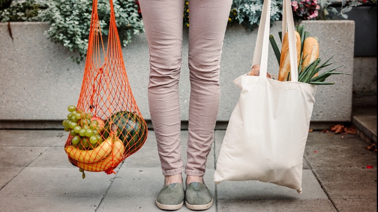 a person holds an orange mesh bag filled with fruits and a white cloth bag with baguettes