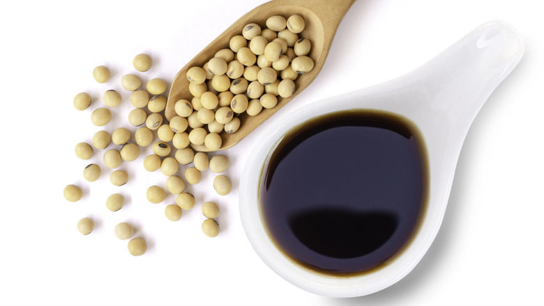 Tasty soy sauce in white ceramic bowl with soy beans in wooden scoop isolated on white background.