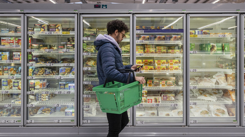 Man checks phone while standing in a supermarket frozen food aisle.
