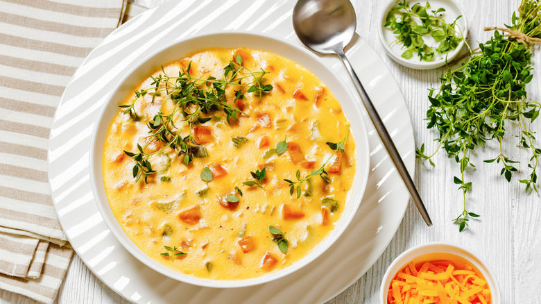 Beer cheese soup in a white bowl next to silver spoon, ramekin of shredded cheese, and herbs