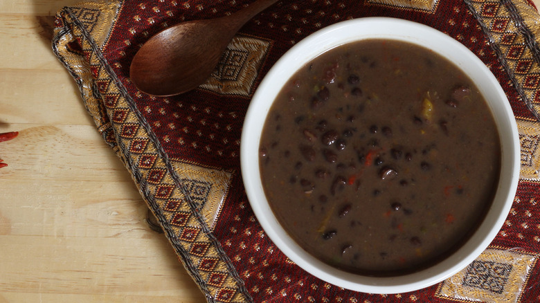 Black bean soup in white bowl next to wooden spoon on patterned place mat