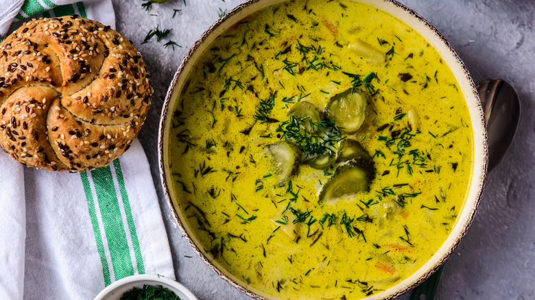 Top down view of light green dill pickle soup in bowl, topped with dill, pickle slices, and next to a slice of bread on tablecloth