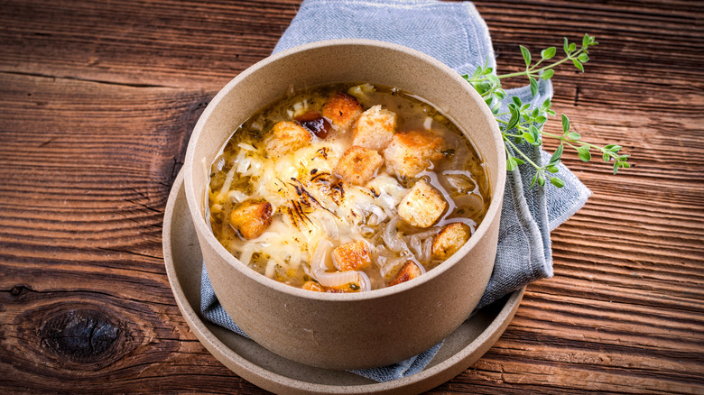 A bowl of French onion soup on a wooden table with blue napkin and herbs for decoration