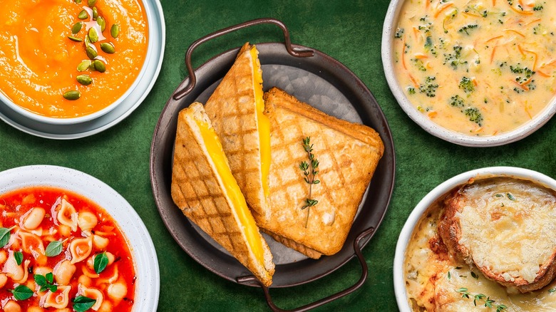 Four soups in white bowls surrounding a cast iron pan with grilled cheese sandwiches