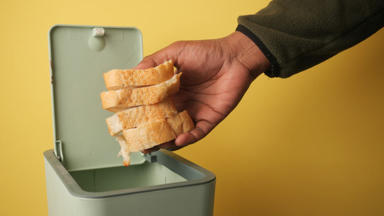 A hand throwing away leftover slices of bread in a green container.