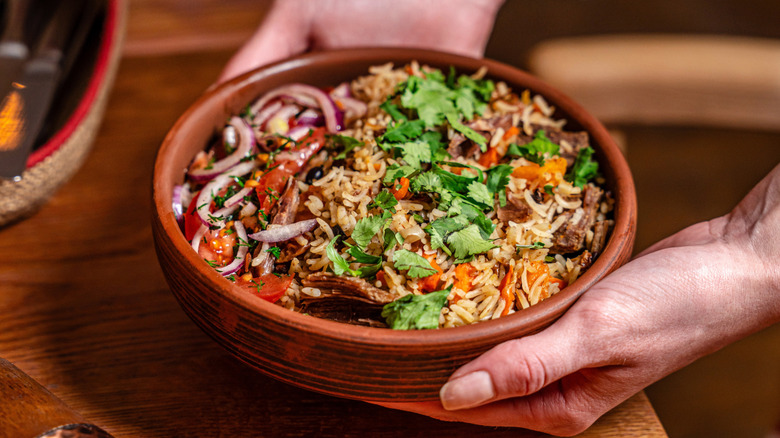 A close-up shot of hands setting a grain bowl with rice, veggies, and meat onto a table