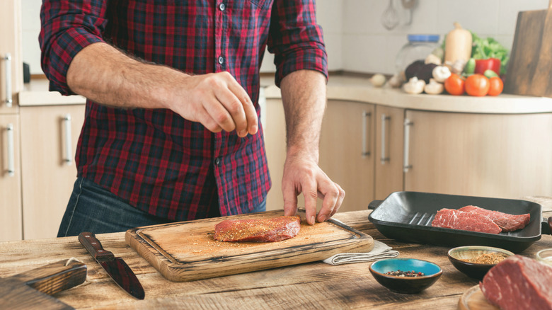 Man's hand dropping seasoning onto cut of beef on cutting board in kitchen