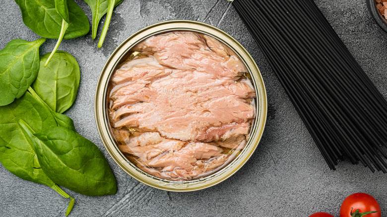 open can of tuna fish by spinach leaves and black sticks on countertop