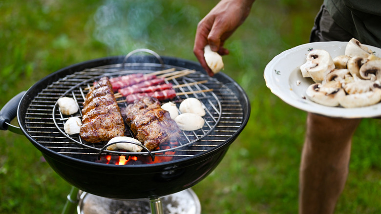 Person placing mushrooms on charcoal grill that already contains beef and chicken