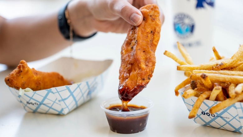 Hand holding a chicken tender that is being dipped in sauce with more tenders and fries in the background