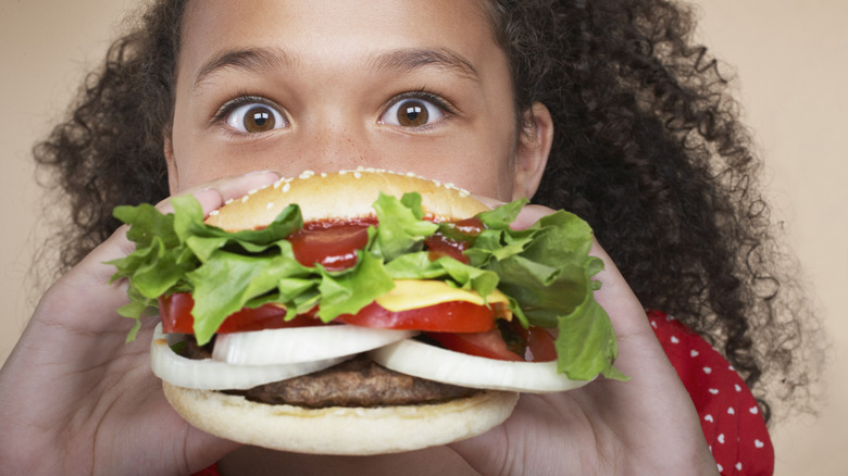 Young girl with eyes stretched wide holding very large burger up to her face