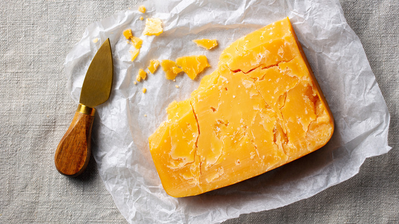 A slice of aged cheddar cheese on piece of wax paper next to a cheese knife.