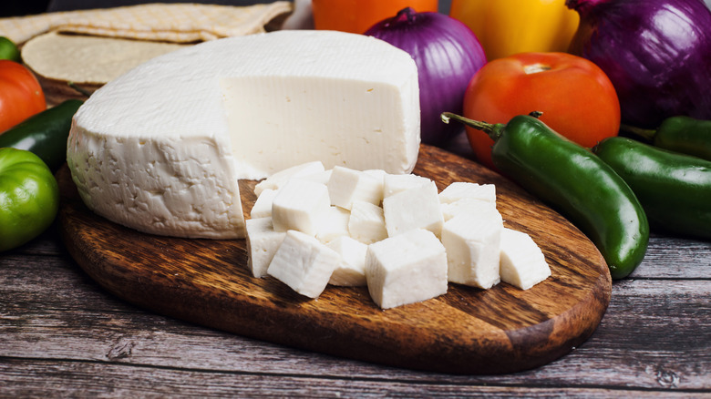 A wheel of Panela cheese partially cut into cubes on a wooden tray in front of various vegetables.