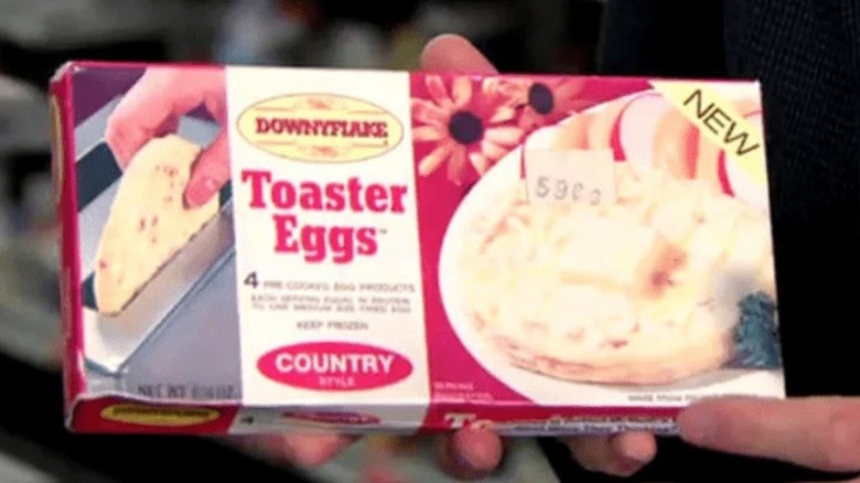 A box of Downyflake Toaster Eggs; a hand pulling a round egg disk out of a toaster on the left; the logo center-left; a plate on a red surface with a toaster egg on it and apple slices on the right