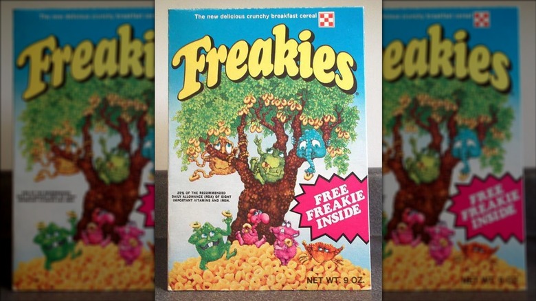 A box of Freakies cereal; a cartoon tree with cereal falling from it with seven monsters; three monsters are in the tree, four are in a pile of cereal on the ground