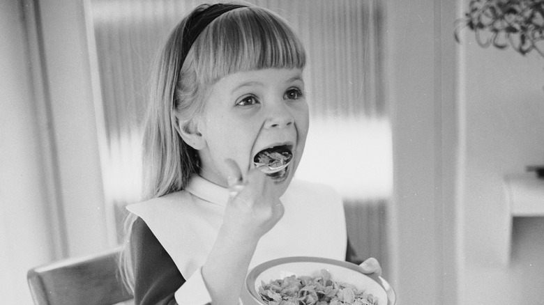 A black-and-white close-up photo of a young blonde girl with long hair and bangs eating cornflakes