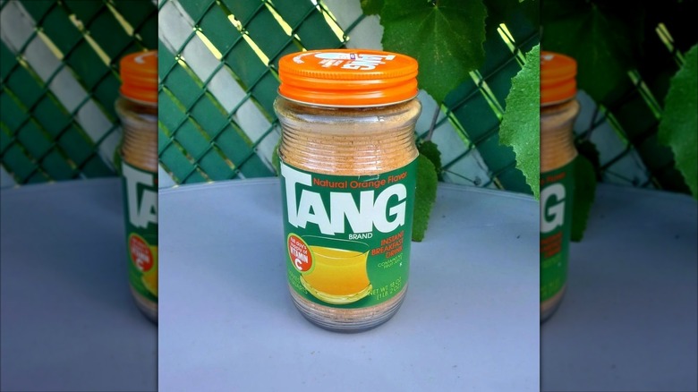 A full vintage jar of Tang sitting on a gray outdoor table with wire fencing and leaves in the background