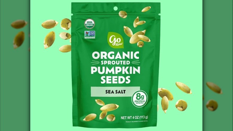 A dark green bag of organic pumpkin seeds sits on a light green background. Pumpkin seeds are on either side of the bag.