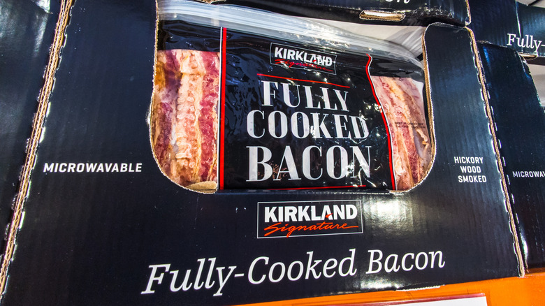 Packages of Kirkland Signature fully cooked bacon rest in a carboard box on a shelf in Costco.