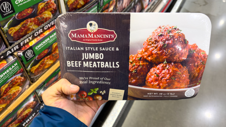 A hand holds a package of Mama Mancini's Italian style sauce and jumbo meatballs. Packages of food are in the background.