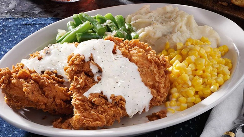 Two pieces of chicken-fried chicken covered in white gravy on a plate with mashed potatoes, greens beans, and corn.