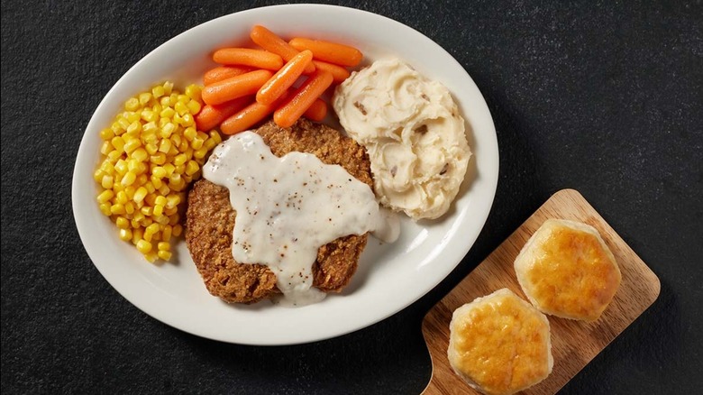 Cracker Barrel country-fried steak covered in gravy on a plate with mashed potatoes, carrots, corn, and two biscuits on a cutting board
