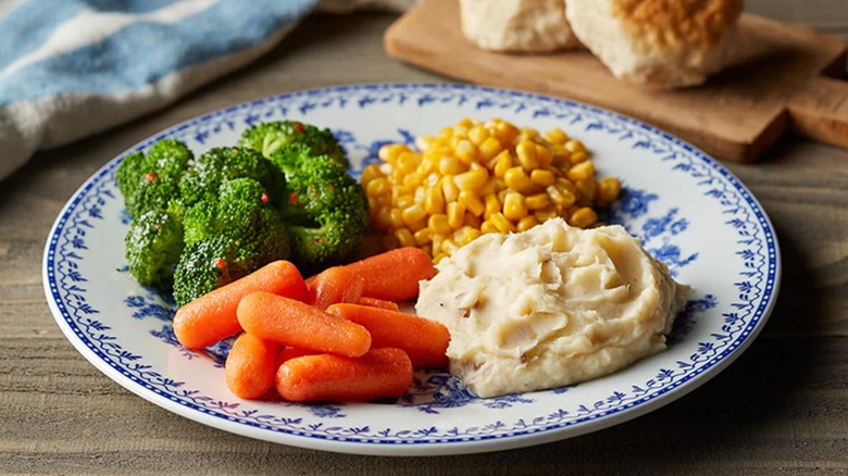 A plate of mashed potatoes, corn, broccoli, and carrots next to two biscuit on a small cutting board.