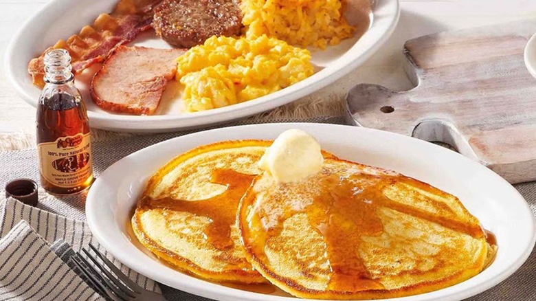 A plate of pancakes with butter and syrup and a plate of grits, ham, bacon, sausage, and hash browns.