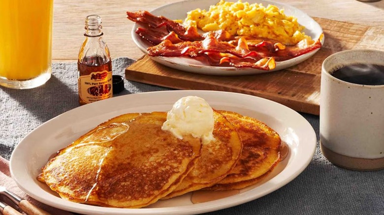 Two pancakes on a plate with butter and syrup next to a plate of scrambled eggs and bacon.