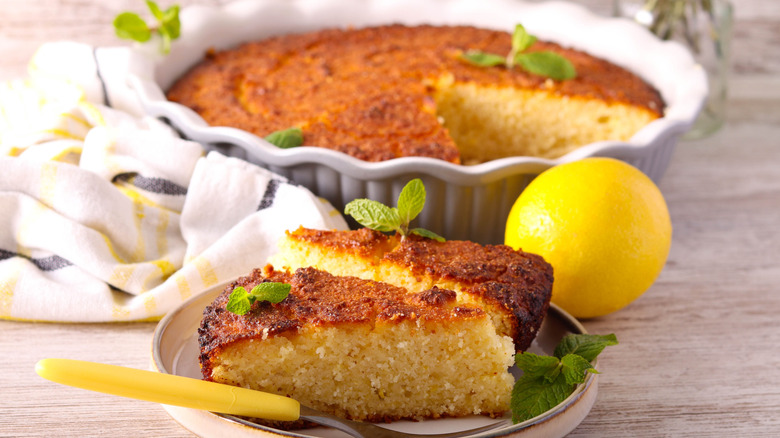Two slices of moist, fresh lemon cake on a white plate with a fork and a partially served whole cake in a ceramic white cake pan garnished with fresh mint and a whole lemon