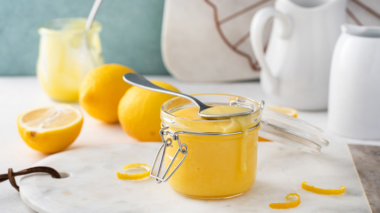 Lemon curd in a jar with a spoon resting on top of it, surrounded by fresh lemons