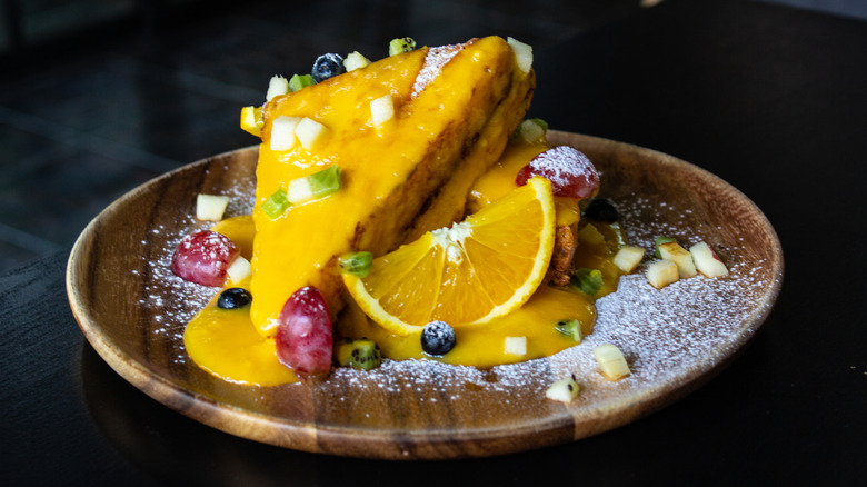 French toast with a citrus-based sauce, garnished with fresh fruit and powdered sugar on a wooden plate