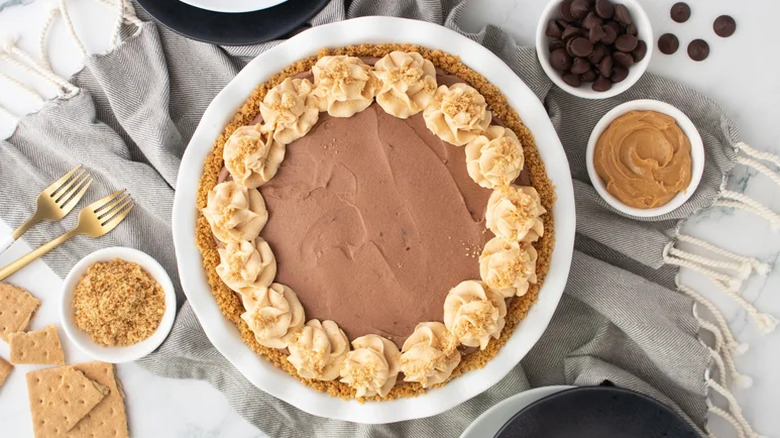Peanut butter pie surrounded by bowls of graham cracker crumbs, chocolate chips, and peanut butter
