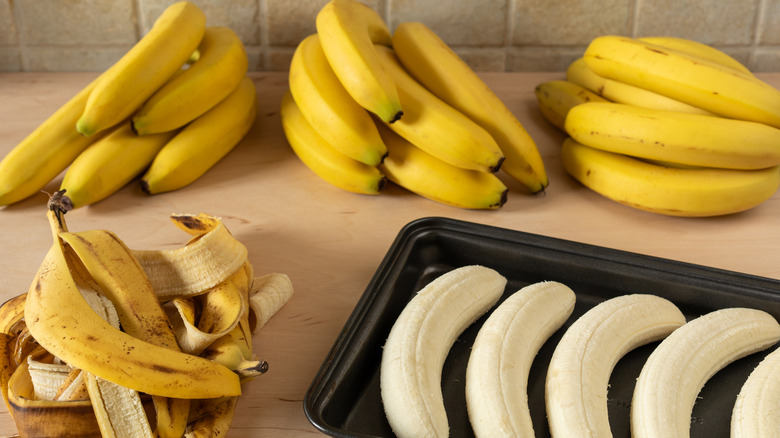 Baking tray with peeled bananas, a heap of banana peels, and three bunches of unpeeled bananas behind
