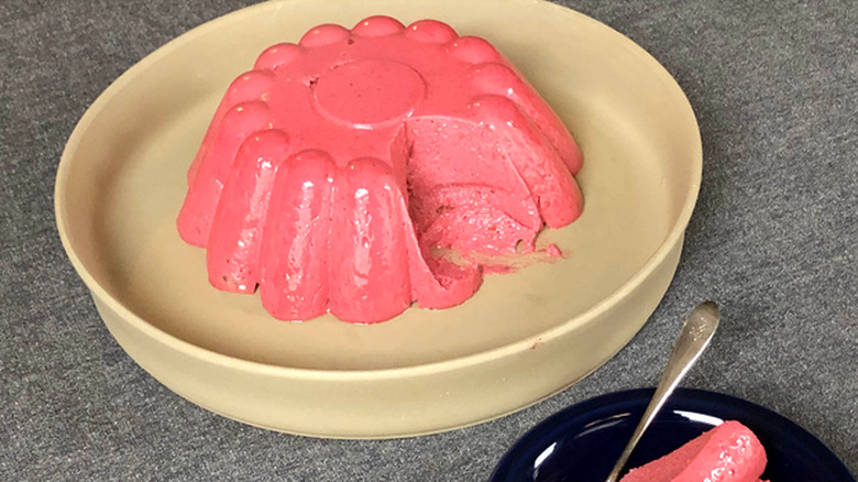 A molded raspberry blancmange on a plate with a slice removed and presented with a spoon on a separate plate